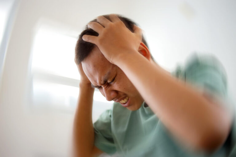 Man grimacing in distress, gripping his head with both hands in a bright room