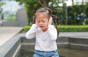 Young girl sits by a fountain with hands over her ears and eyes closed, wearing a white lace top and blue jeans.