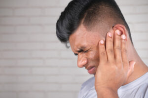 Man with short hair holding his ear and wincing in pain, possibly an earache or headache, against a light brick wall.