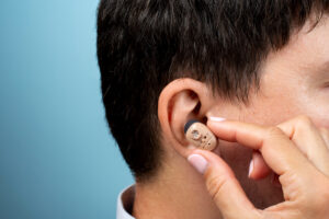 Close-up of a person inserting a beige behind-the-ear hearing aid into their ear against a blue background.